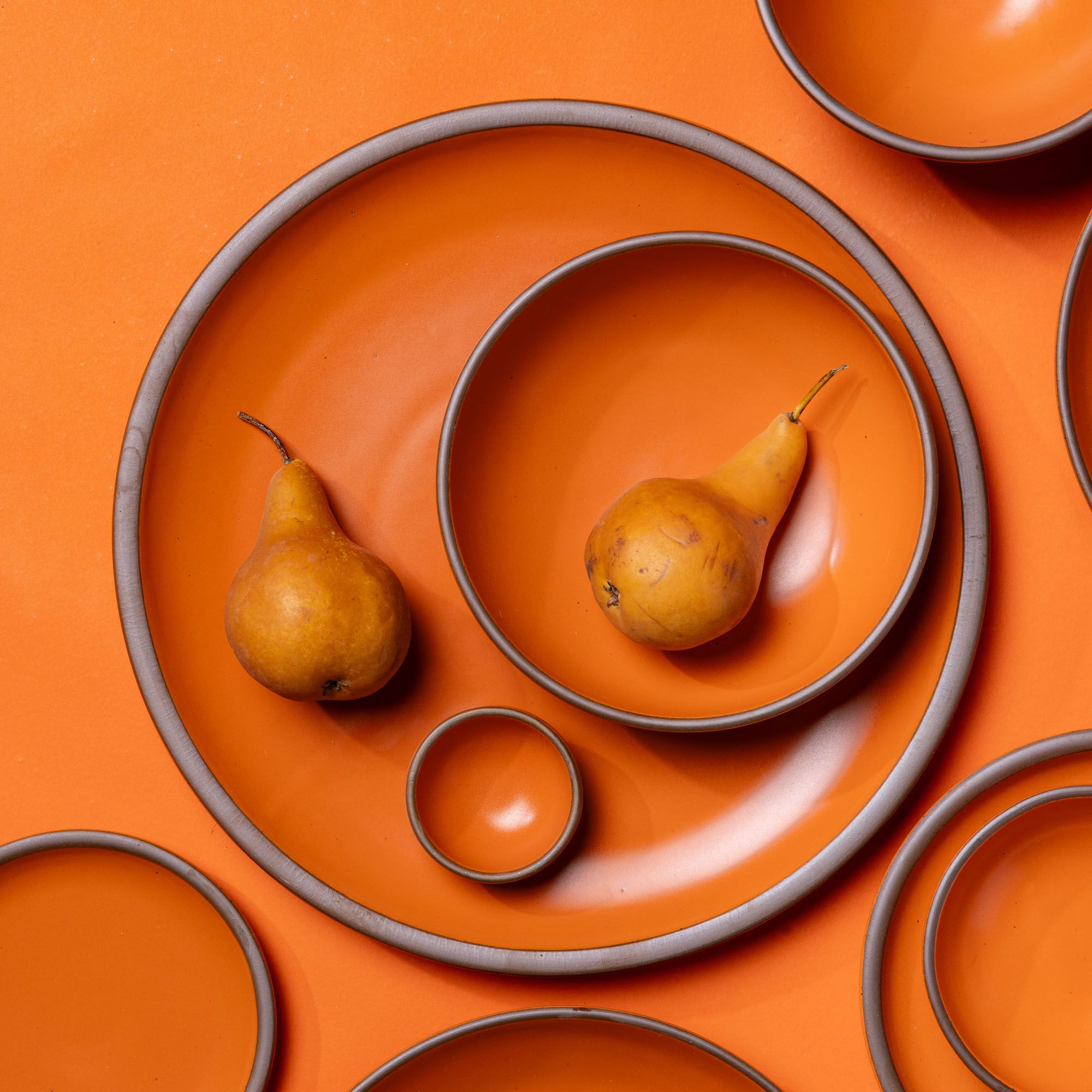 An artful arrangement of a large ceramic platter in a bold orange color with a smaller plate with pears on each.