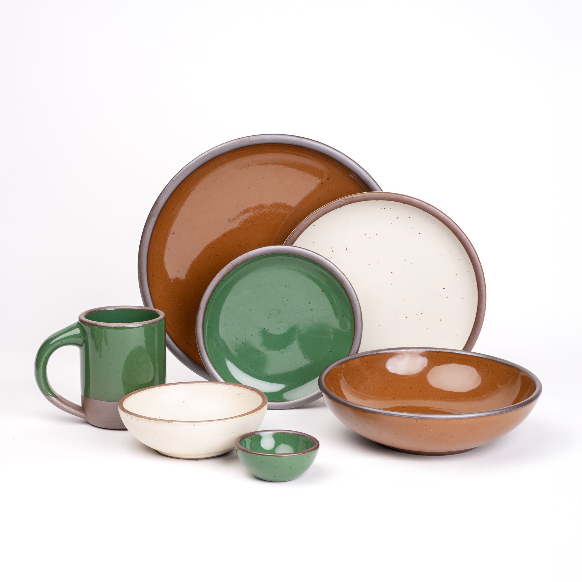 The Mug, bitty bowl, and cake plate in a glossy emerald green, everyday bowl and dinner plate in a glossy caramel brown, a side plate and breakfast bowl in a white cream paired together featuring iron speckles