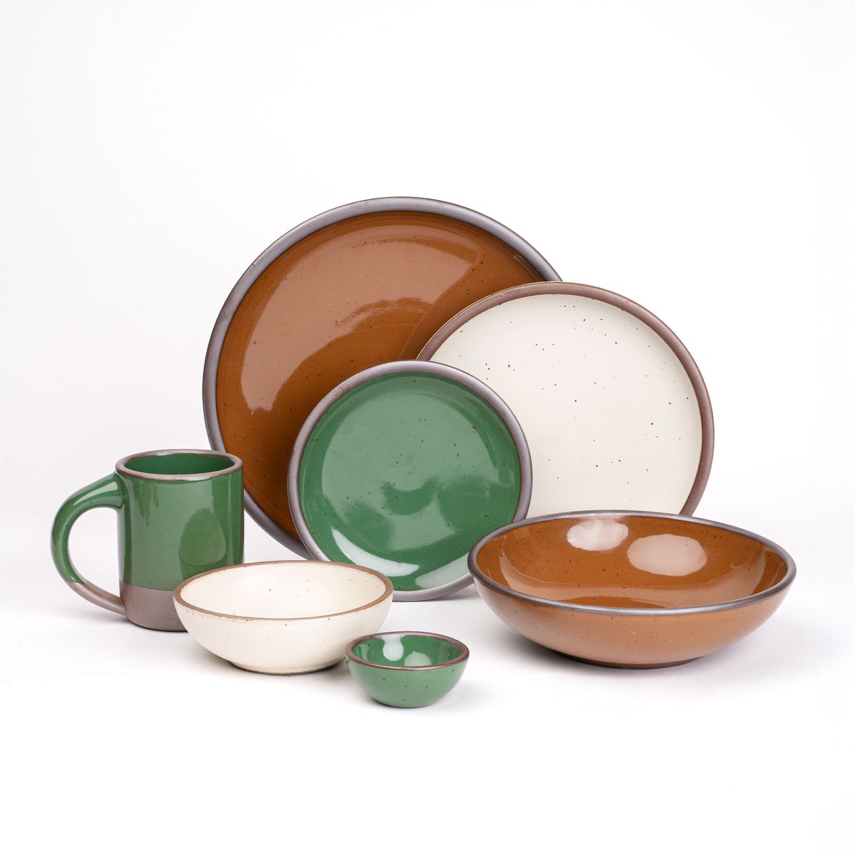 The Mug, bitty bowl, and cake plate in a glossy emerald green, everyday bowl and dinner plate in a glossy caramel brown, a side plate and breakfast bowl in a white cream paired together featuring iron speckles