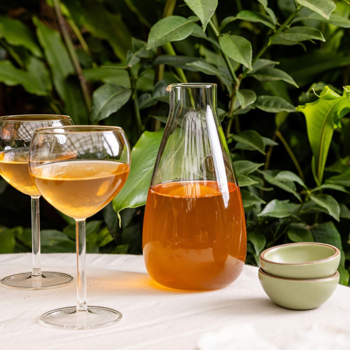 On a table outdoors, sits 2 simple wine glasses with a sophisticated tall glass wine carafe, filled with orange wine. To the right is 2 stacked tiny bowls in a sage green color.