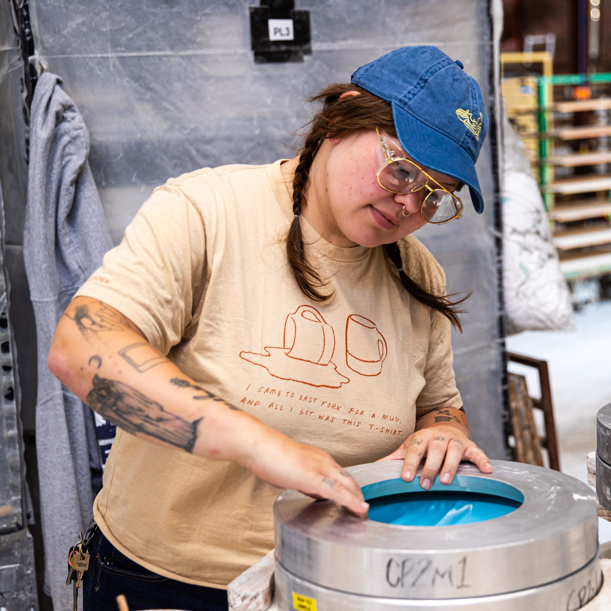 In a manufacturing setting, a person is wearing a classic blue baseball hat with yellow embroidery on the front depicting a feast of fish, fruit and wine.