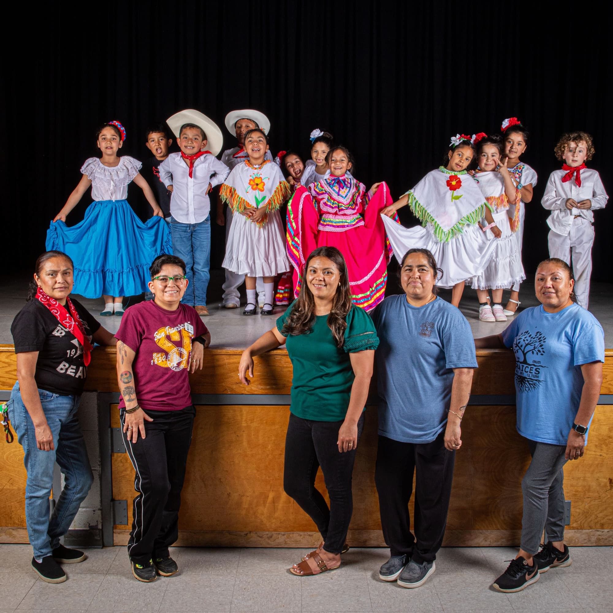 On a stage are a bunch of young kids dressed up and smiling with colorful skirts, white button down shirts, and cowboy hats or flowers. In front of the stage are five adults smiling and staring at the camera.