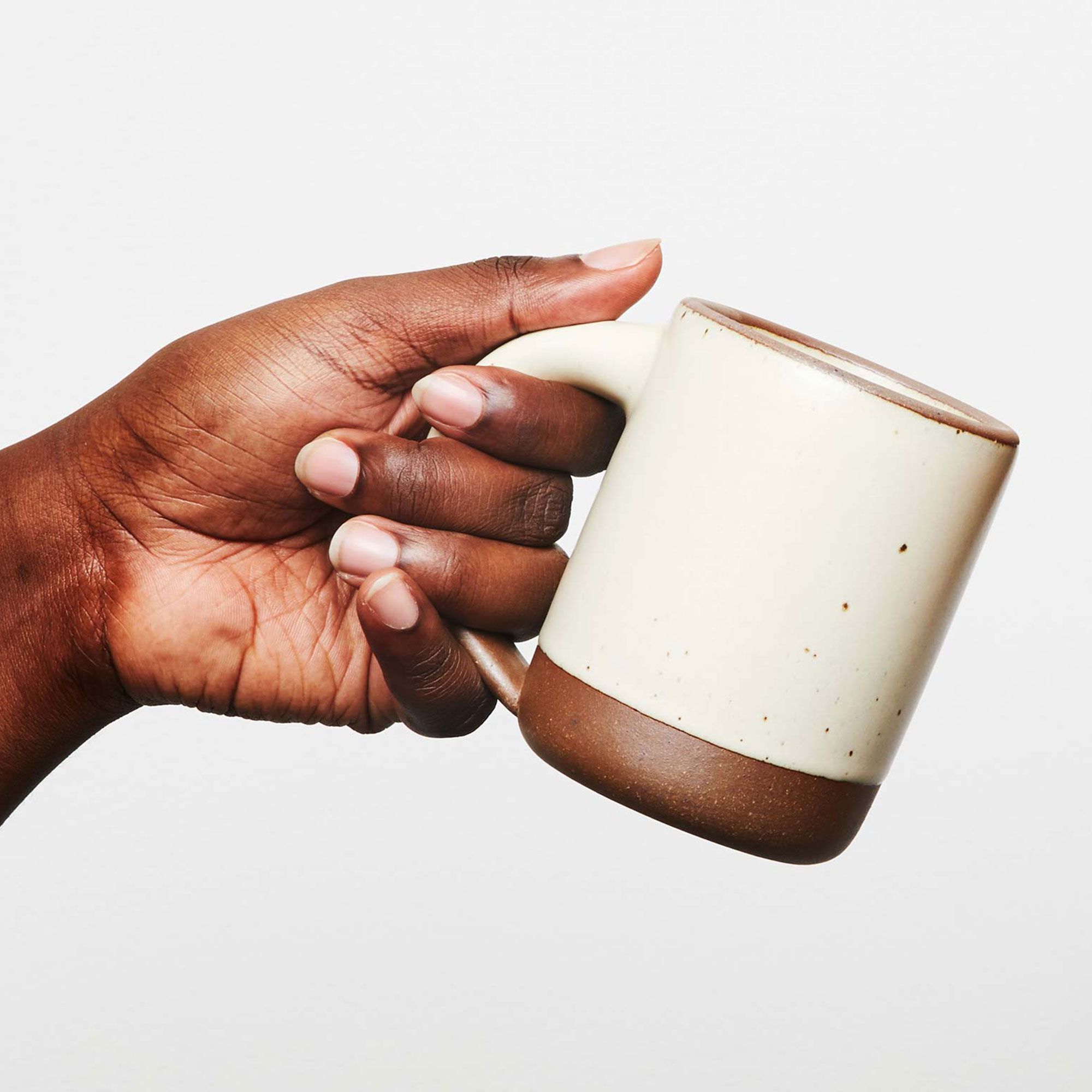 A hand holding a medium sized ceramic mug with handle in a warm, tan-toned, off-white color featuring iron speckles and unglazed rim and bottom base.