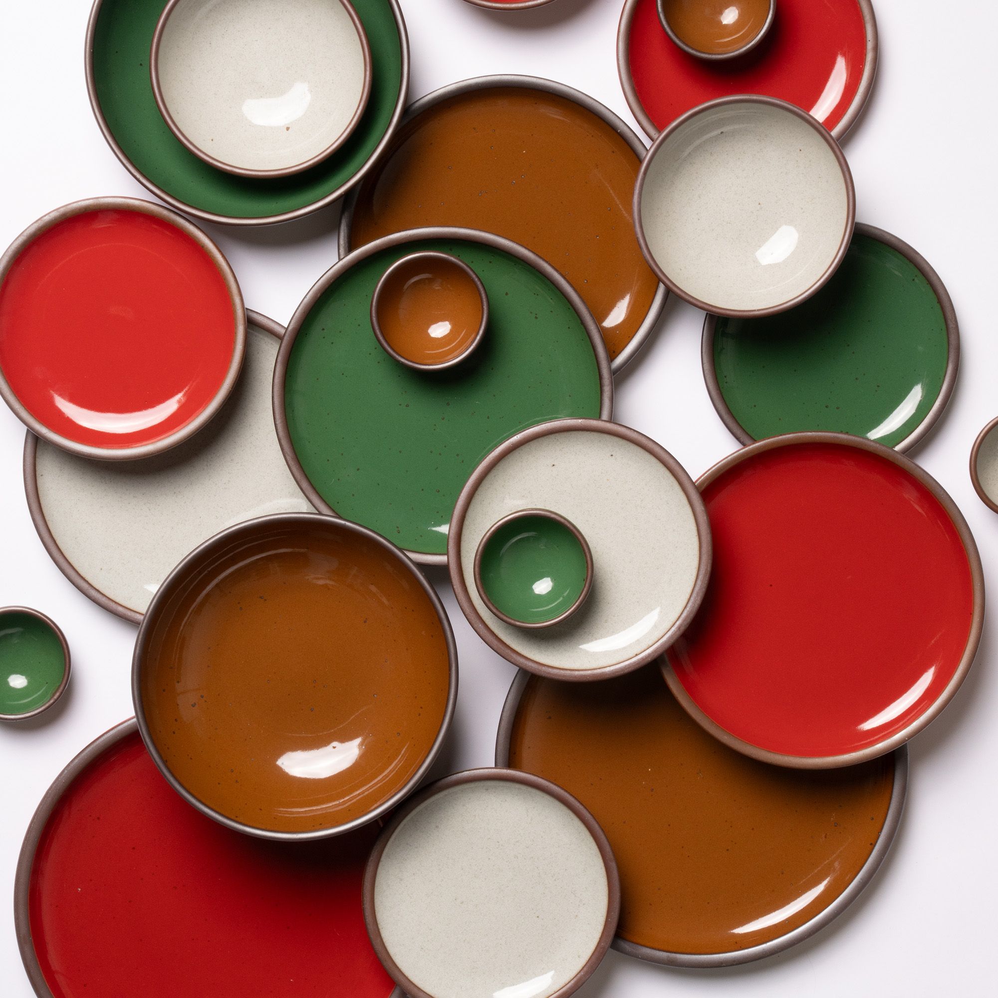 An overhead view of ceramic plates and bowls in various sizes, in a glossy red, green, brown, and icy white colors.