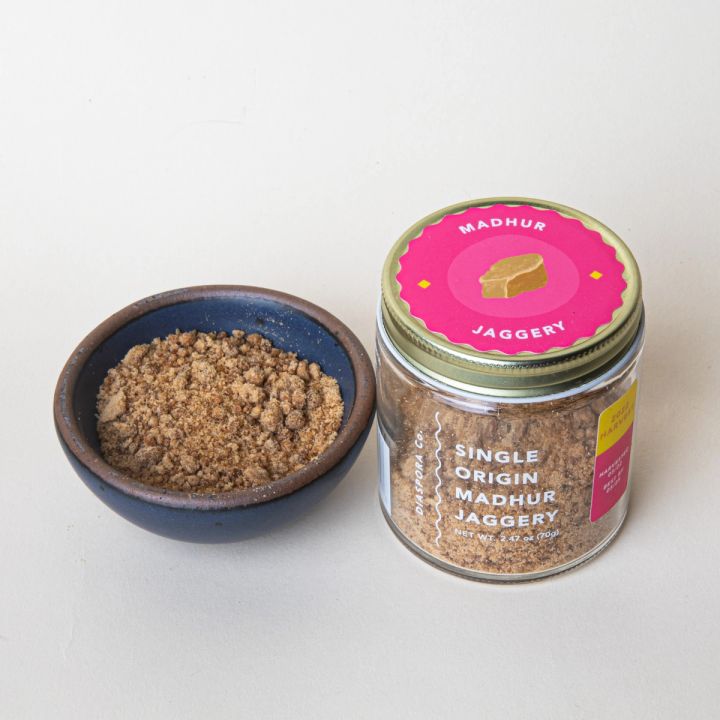 Short glass jar with a metal screw top with a pink label, the jar reads "Single Origin Madhur Jaggery", with a bitty bowl of jaggery next to it.