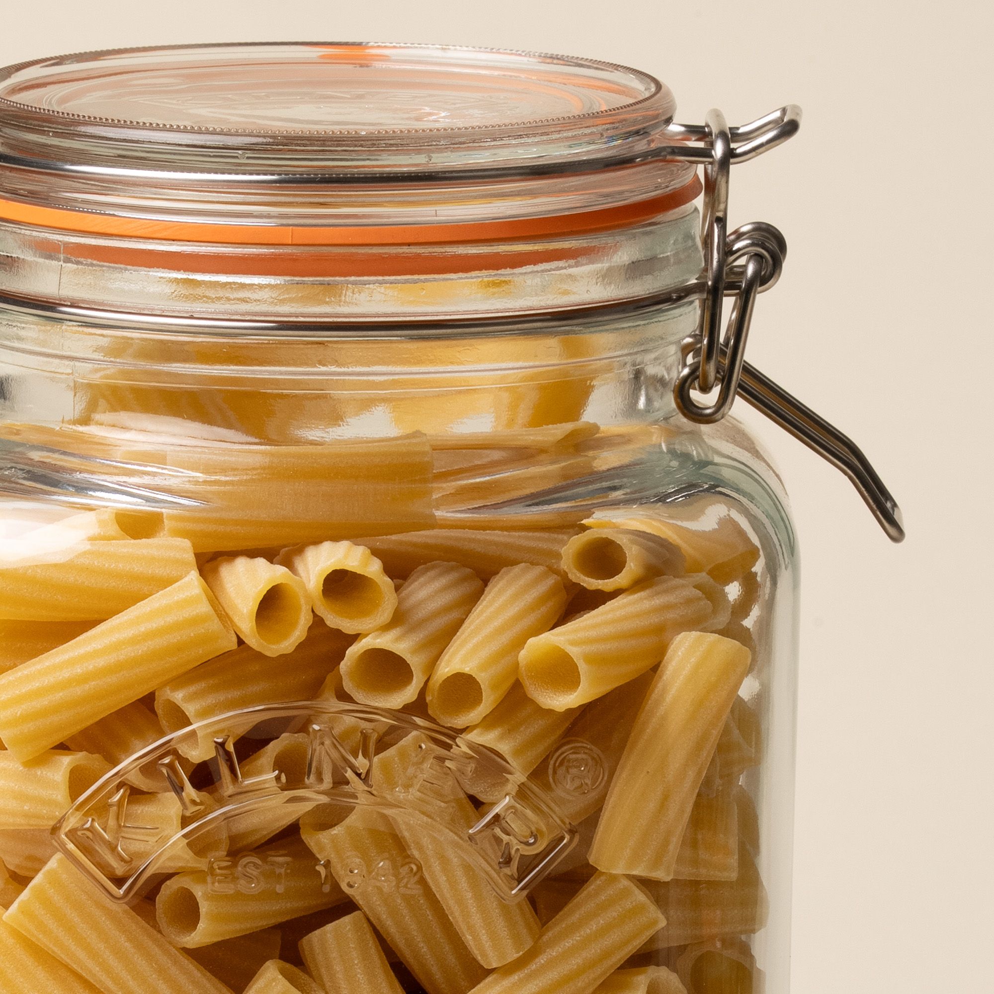 A close up of a storage glass square jar filled with dried pasta, topped with a lid with a rubber seal and steel clip closure.