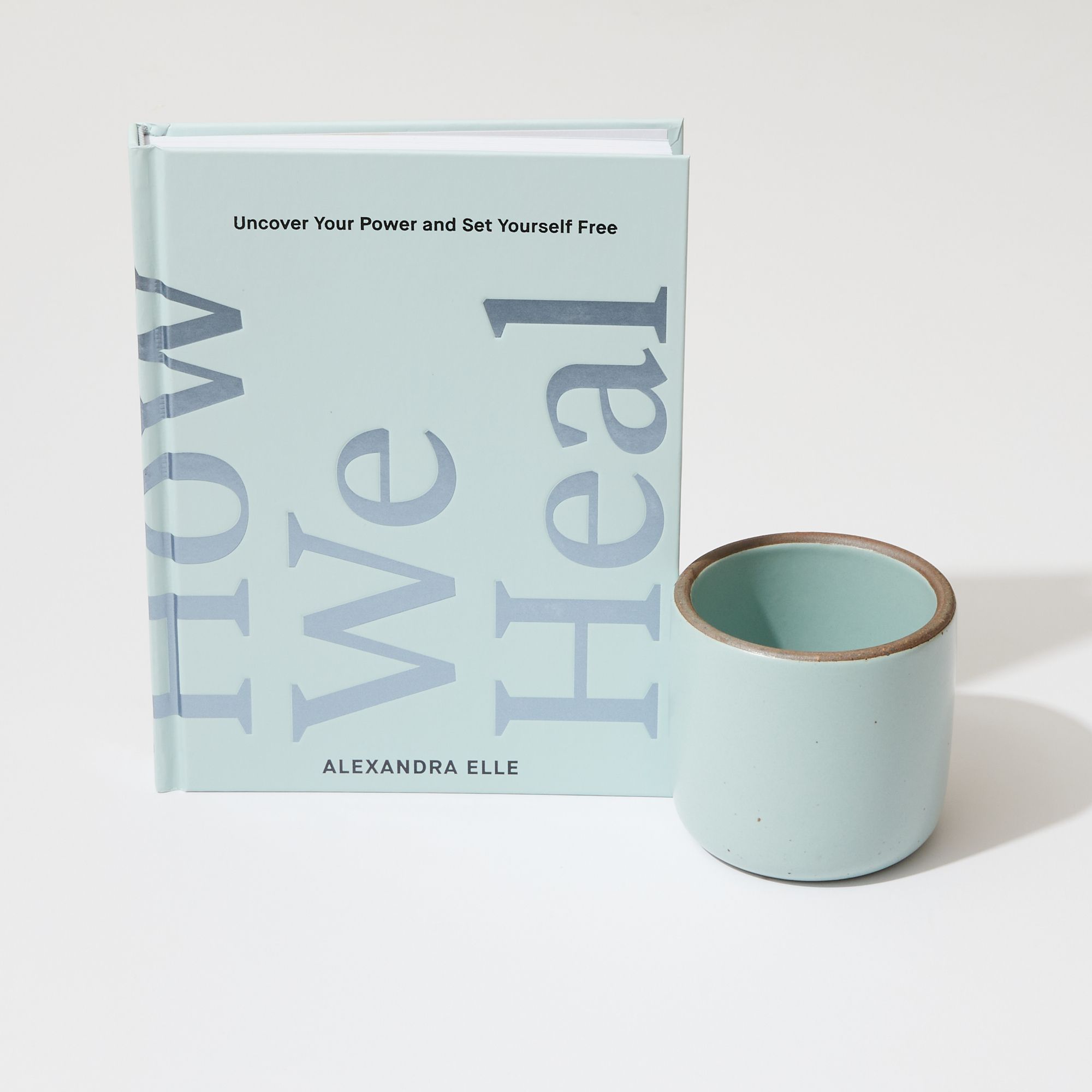 A pale turquoise book standing up that reads "How We Heal", with a ceramic cup in a pale turquoise color next to it. 