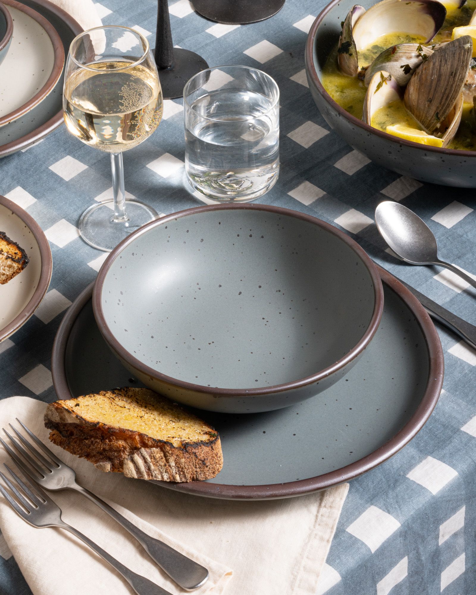 Medium shallow bowl and dinner plate in a blue-grey color, with silverware on a blue-grey crosshatch tablecloth