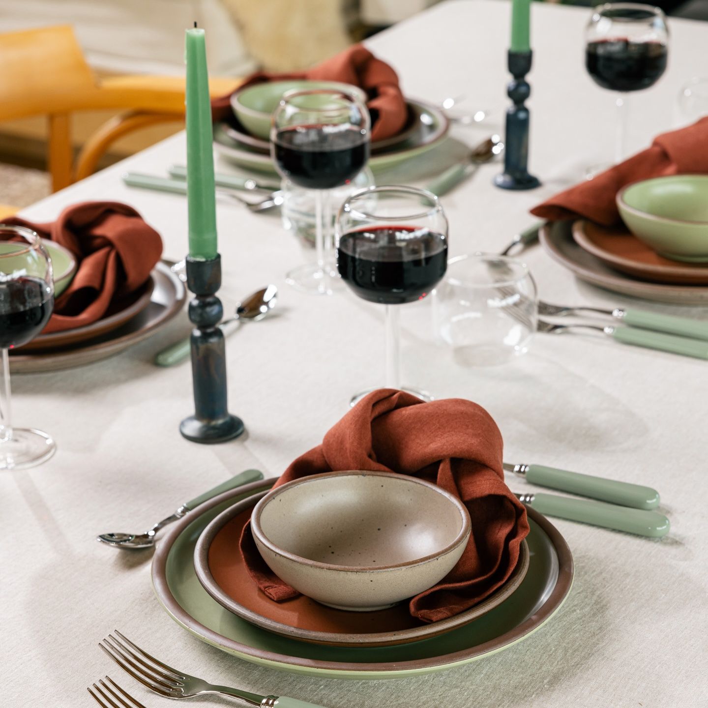 A table filled with several place settings, featuring stacked plates and a bowl in sage green, terracotta, and natural colors, a chic folded terracotta napkin, sage flatware, wine glasses, and green taper candles in iron candleholders.
