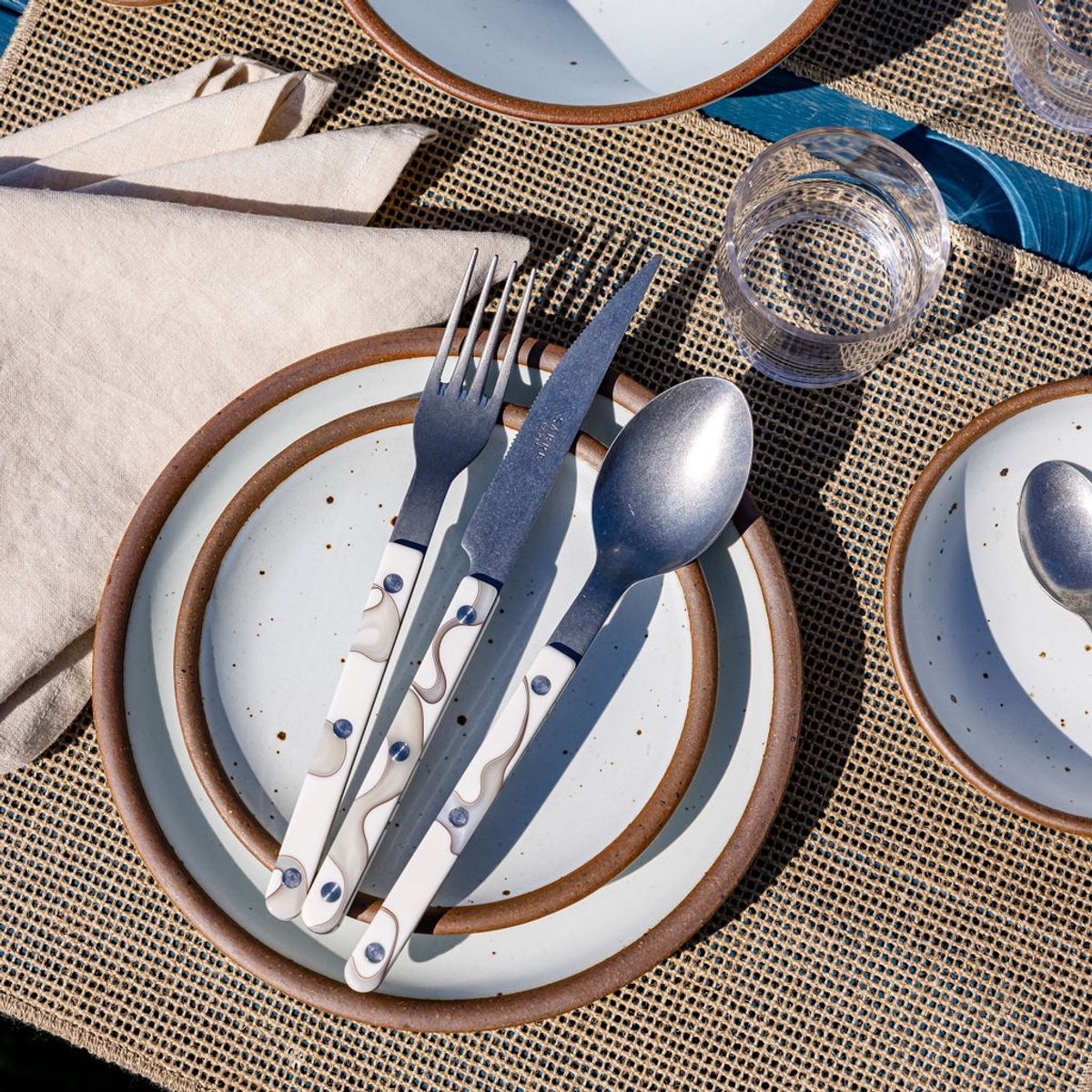 A table setting with a neutral woven placemat, stacked cool white ceramic plates, and ivory marble-handled fork, knife, and spoon.