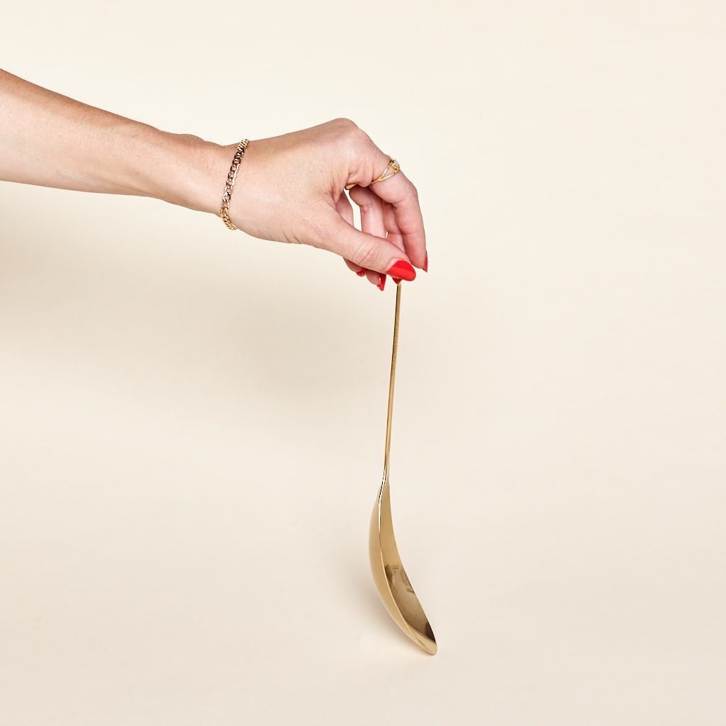 A forearm and hand at top and fingers hold the top of the handle of a brass spoon