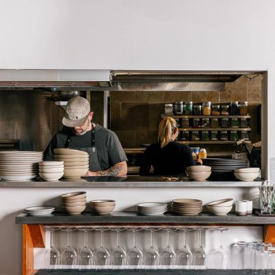 In a restaurant setting, there is a kitchen window with chefs plating food and a lineup of ceramic bowls and plates along with wine glasses below.