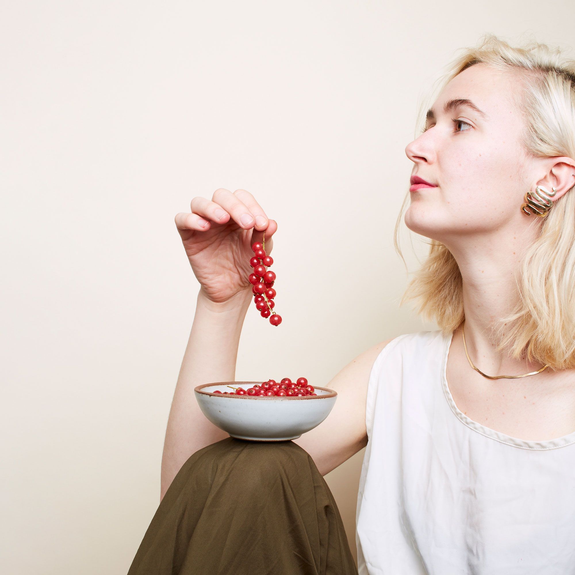 A person with blonde hair has a shallow ceramic bowl in a light gray color resting on their knee and is holding a stem of berries above it.