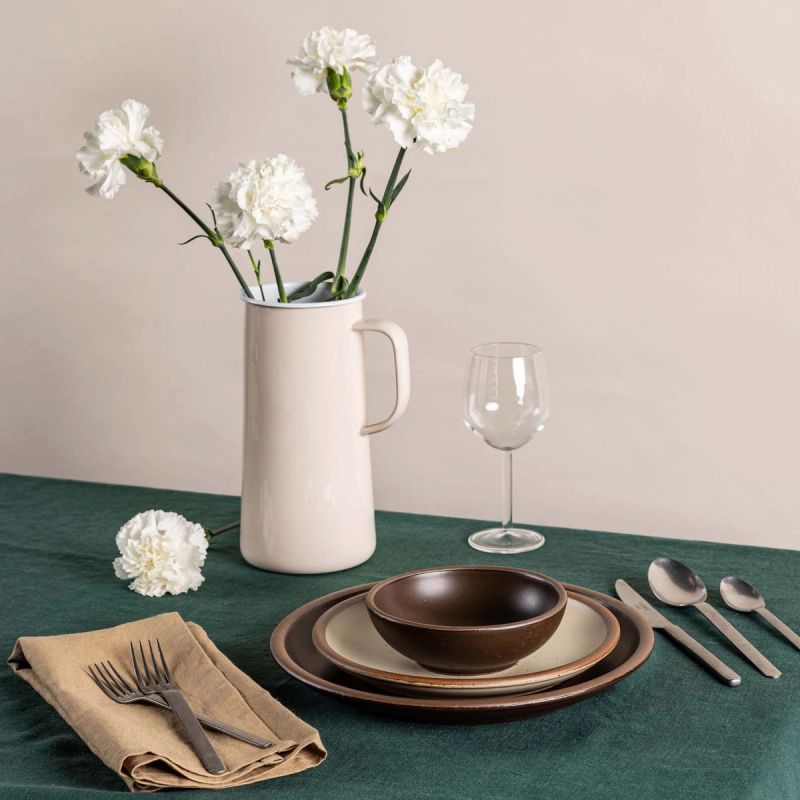 On a table sits a simple place setting with brown and off-white plates and a bowl, steel flatware, a wine glass, and an enamel pitcher in cream with flowers inside.