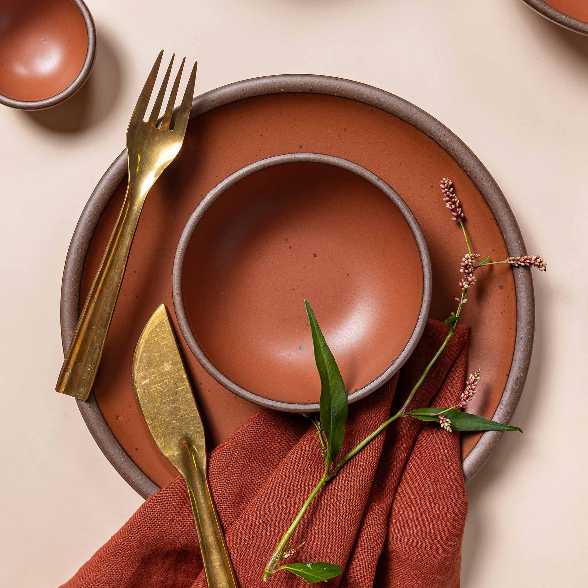 A ceramic dinner plate and small shallow bowl in a burnt terracotta color with a brass flatware, napkin, and greenery.