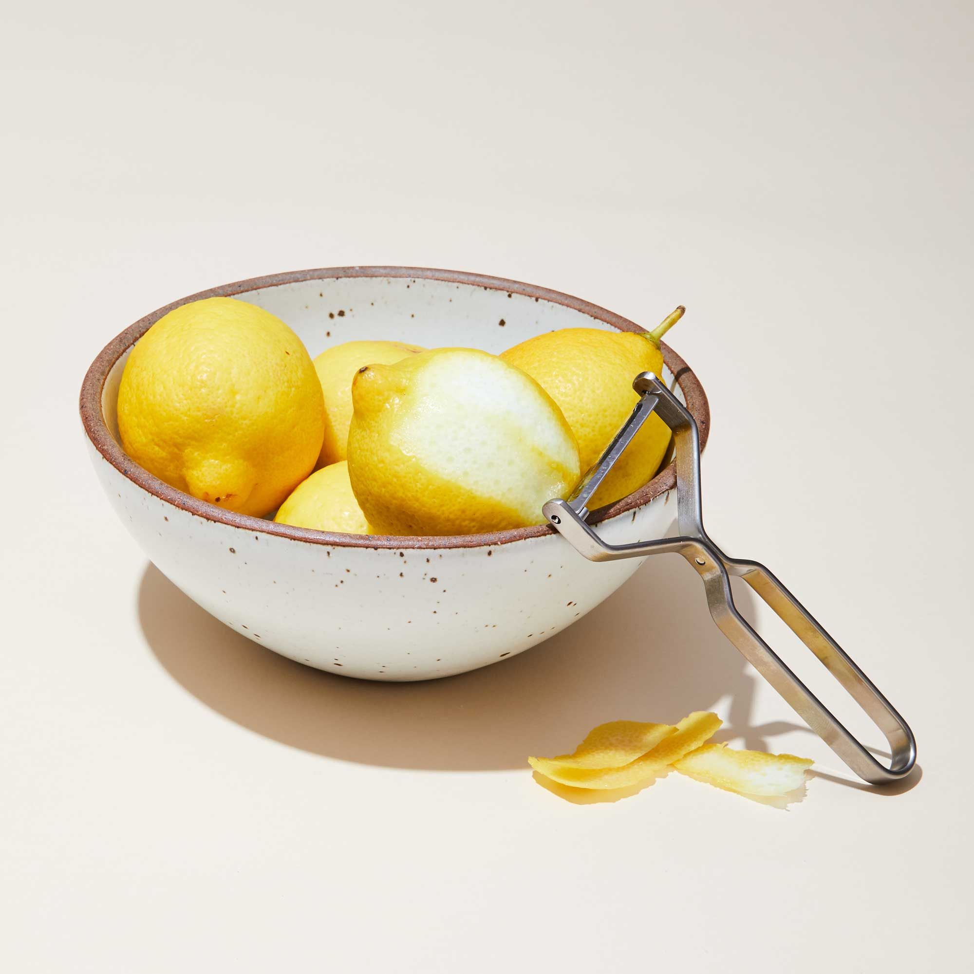 A stainless steel peeler rests against the rim of an East Fork bowl that is filled with lemons, one of which is partially peeler.
