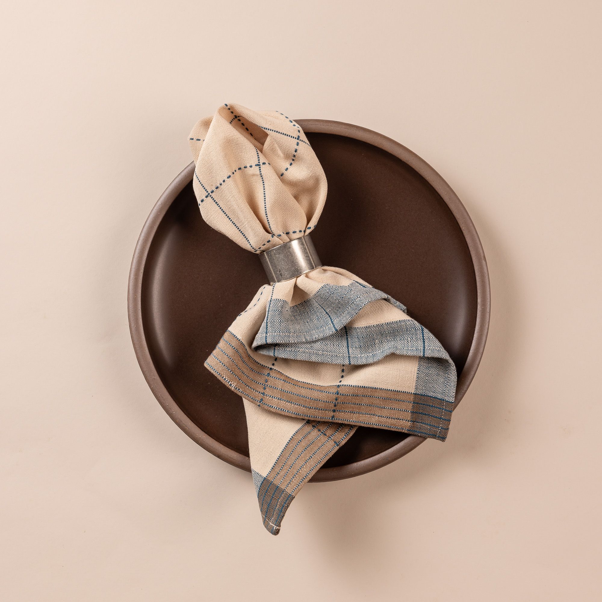 A natural folded napkin pulled through a pewter napkin ring sits on a deep brown dinner plate. The napkin is designed with turquoise gridlines with light blue and tan striped edging.