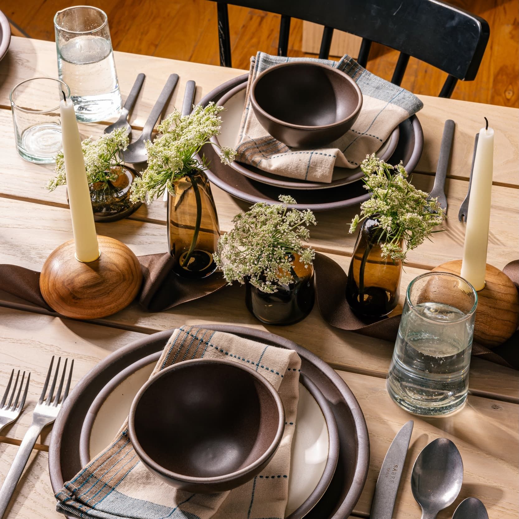 On a set table is two place settings with dark brown and cream ceramic plates and bowls, simple water glasses, steel flatware, simple filler flowers in vases, and round short wood candleholders with cream taper candles.