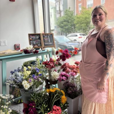 A blonde woman with tattoos and a pink apron standing in front of flowers with a window behind her.