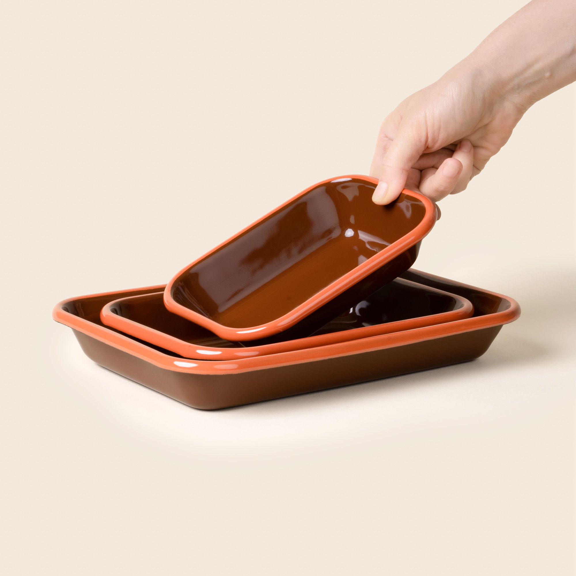 A hand placing a small dish into a nesting trio rectangular baking dishes in a dark red-brown color with an orange rim.