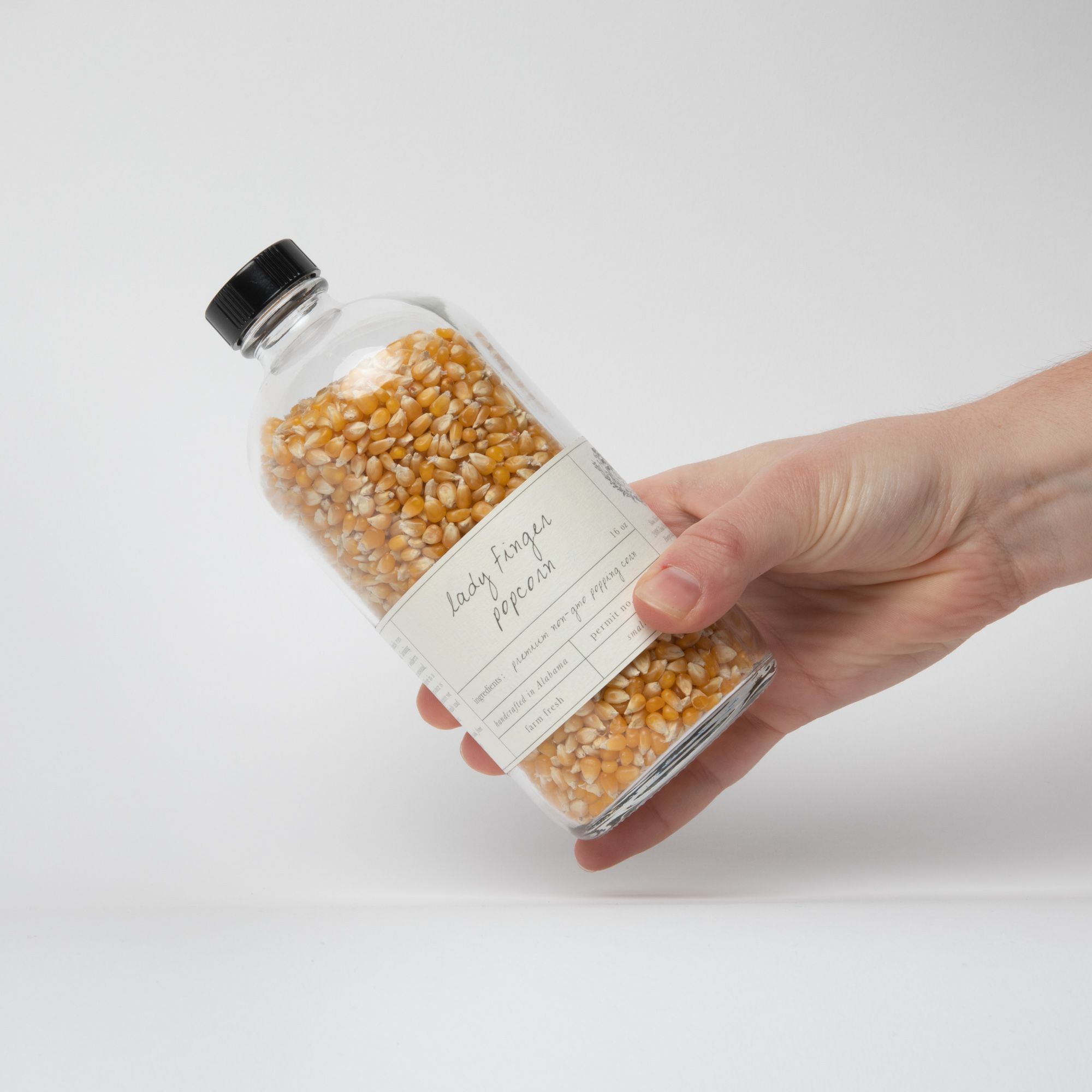 Hand holding a glass bottle of popcorn kernels with an off white label that reads "Lady finger popcorn".