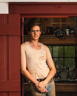 John standing and leaning on the doorway of his small white cabin pottery studio.