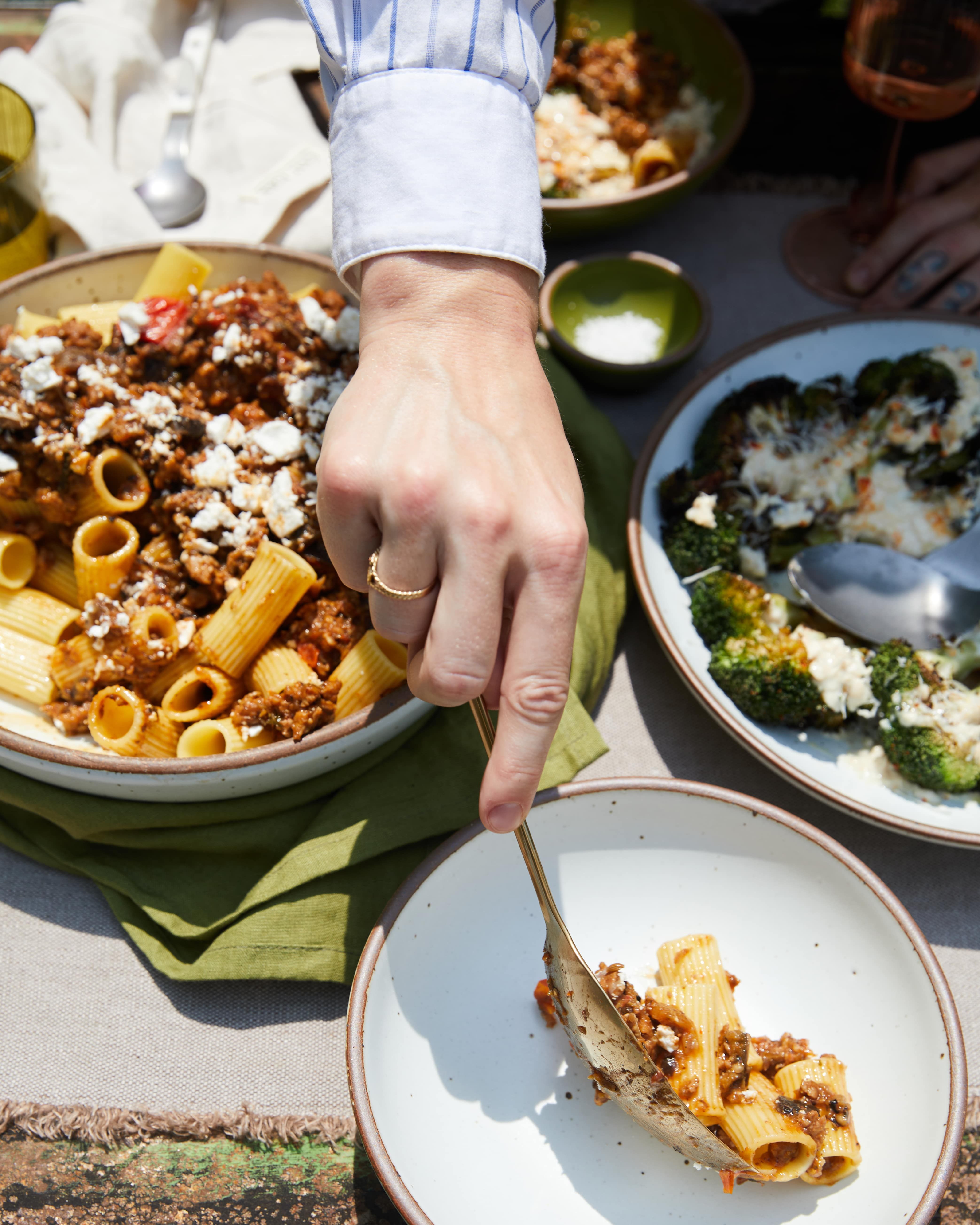 A hand plates rigatoni from the large serving bowl to a white ceramic plate