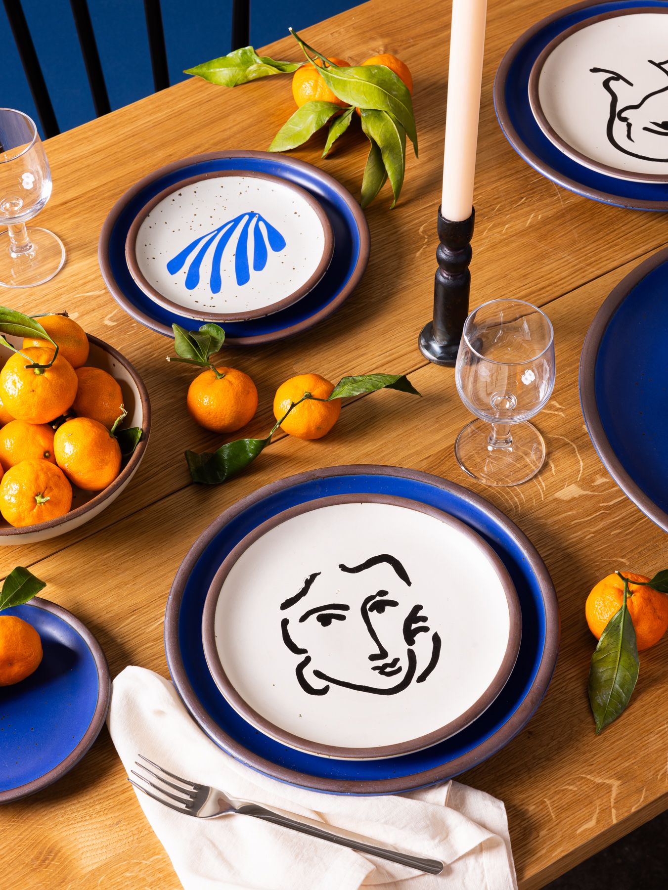 An overhead view of ceramic plates and mug featuring blue decals of artwork by Henri Matisse, along with plates in a cobalt blue color. There is also a red tablecloth, silverware, flowers, and oranges.