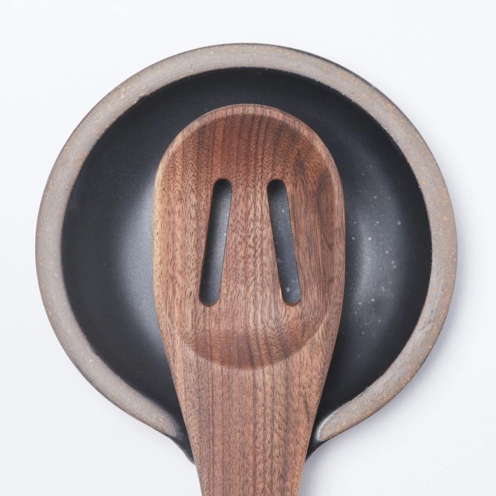 A wooden slotted spoon sits on a circular ceramic spoon rest in a graphite black color featuring iron speckles and unglazed ridge rim