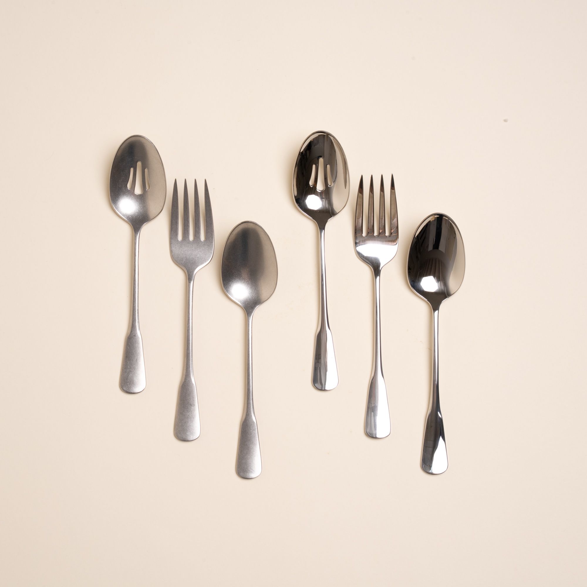 Two sets of stainless steel serving utensils including a serving fork, serving spoon, and pierced spoon. One is in a polished finish, the other in a tumbled matte finish, both a classic design