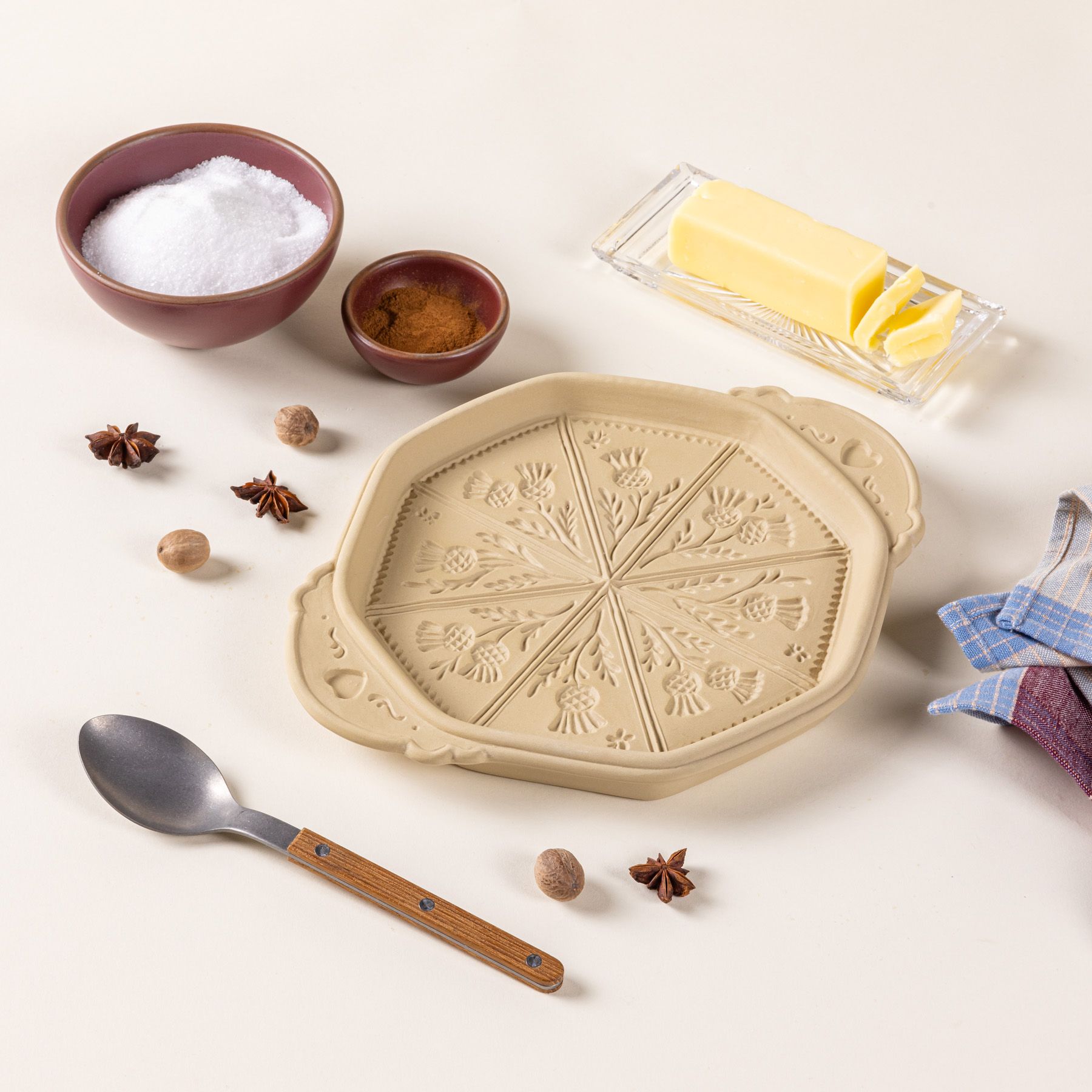 A shortbread pan with a thistle flower design on the bottom, with other baking tools like a spoon, napkin, butter dish, and little bowls of sugar and cinnamon