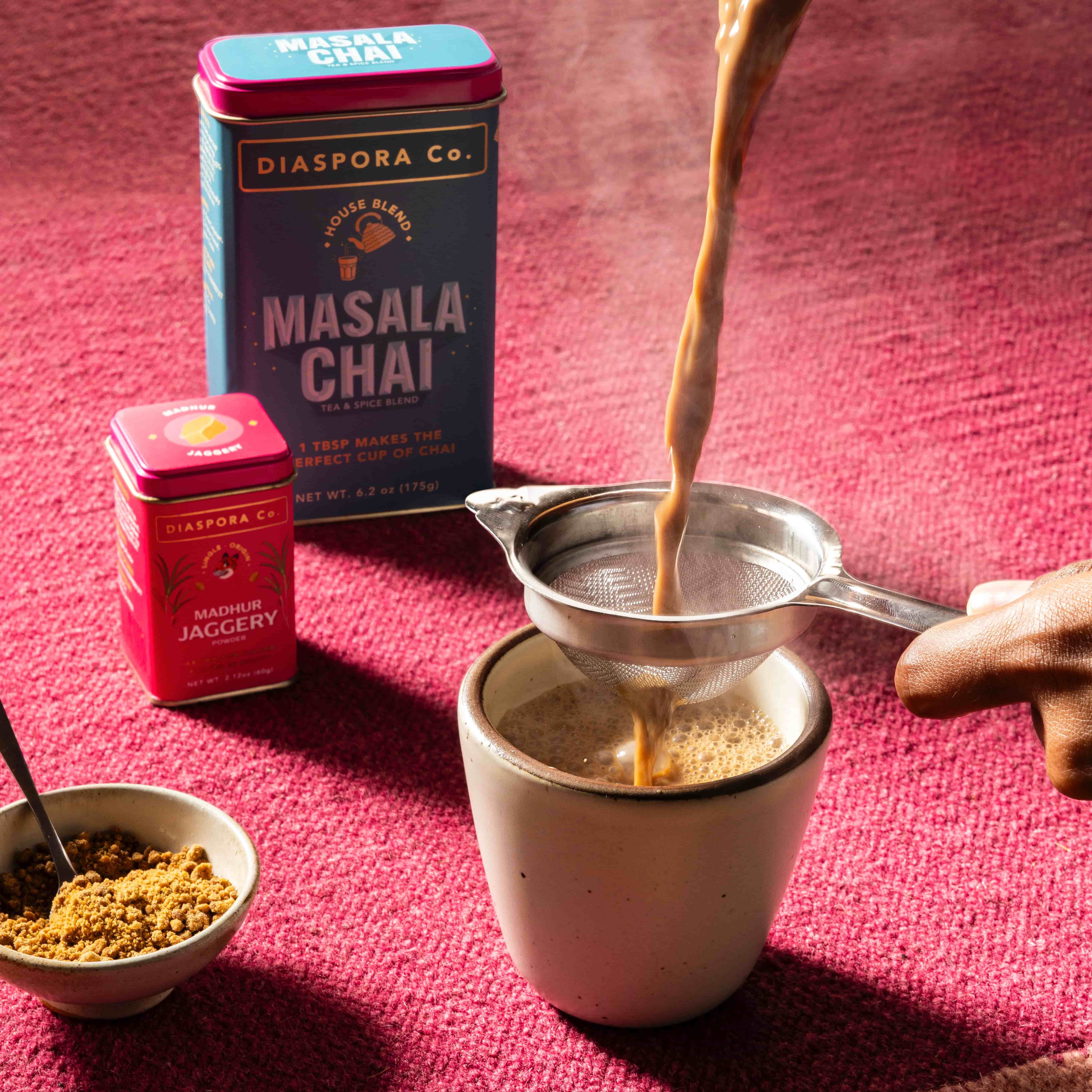 A hand holds a strainer over a small ceramic cup pouring chai, next to two food tins of masala chai and jaggery, a small bowl of jaggery, all against a bright pink background.