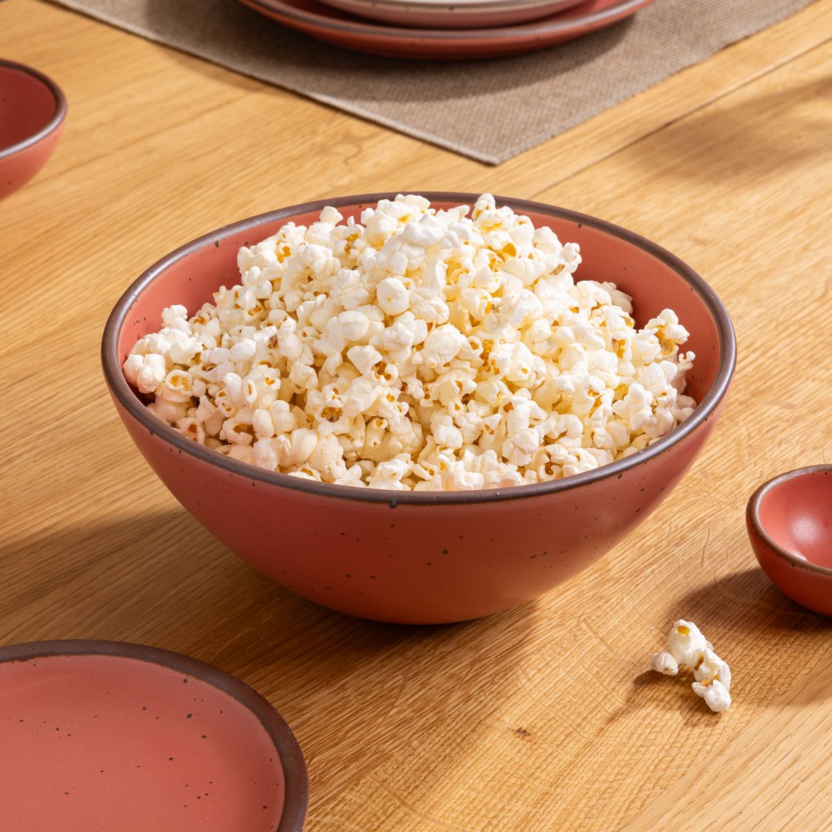 Medium-sized watermelon pink round ceramic bowl filled with popcorn on a wooden table set with matching dishware and a woven placemat.