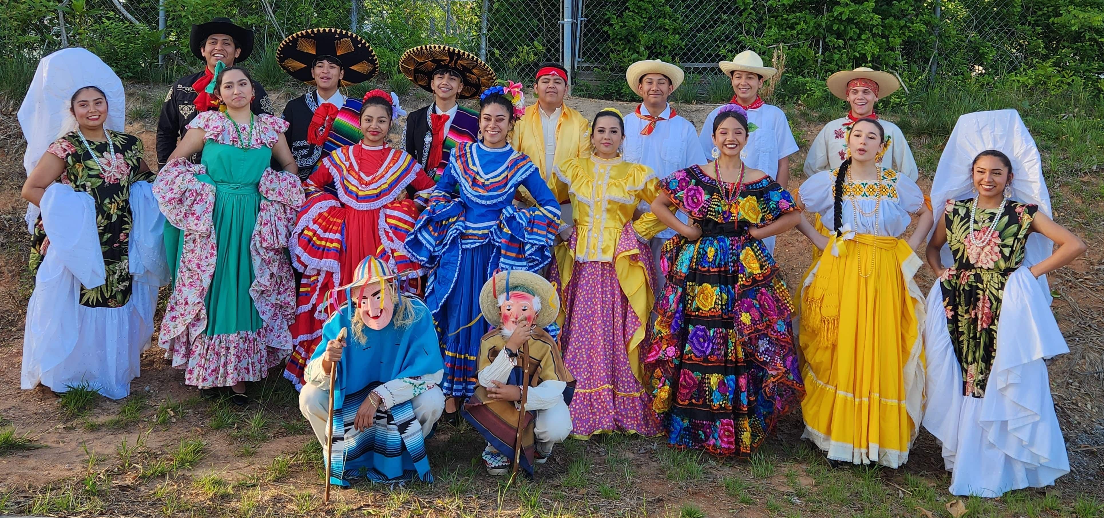 Dancers from the Ballet Folklórico Raíces are grouped together for a photo outside. They are smiling and wearing colorful folkloric dresses, suits, and costumes.