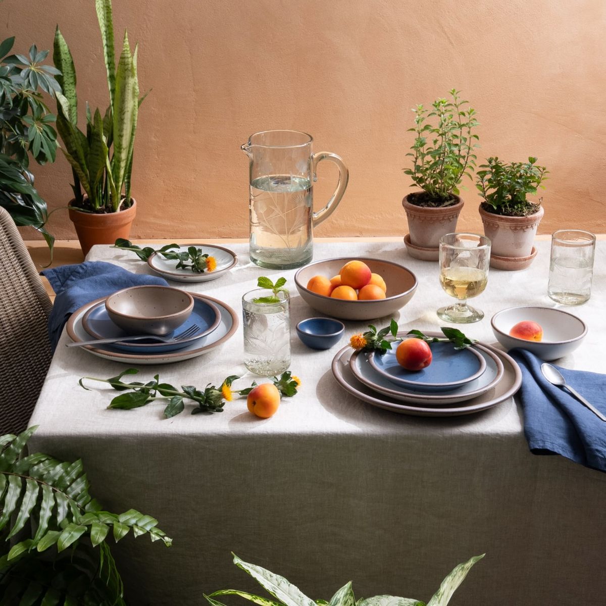 Summer table set with stoneware dishes, blue napkins, fruit, fresh herbs, and glassware, surrounded by potted plants.