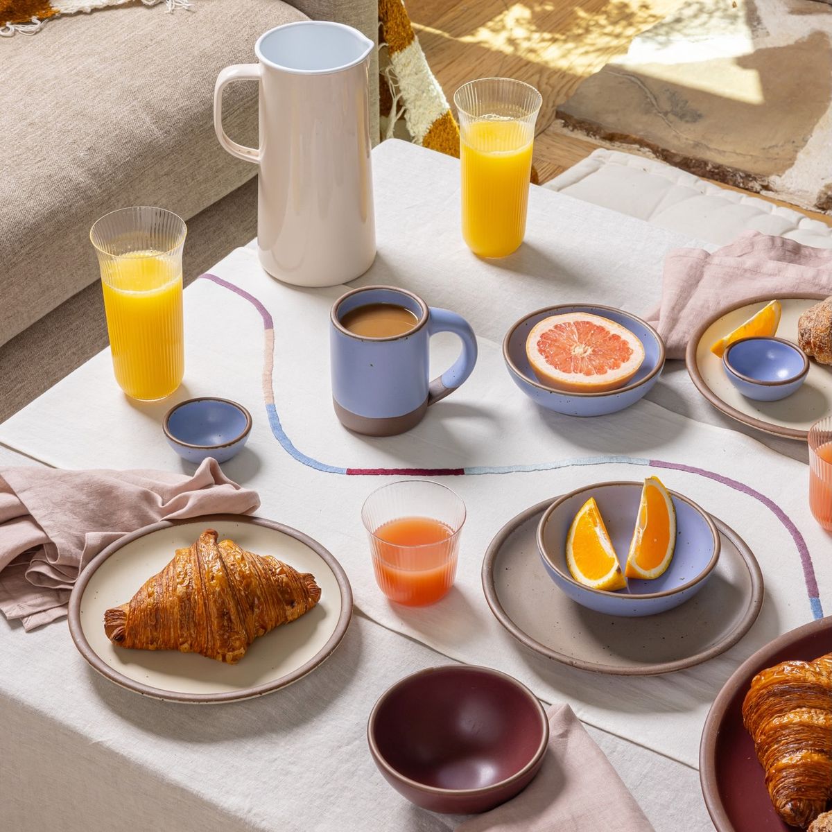 A large coffee table with a runner with a thin embroidered line curving down the runner, and a casual breakfast set-up on the table