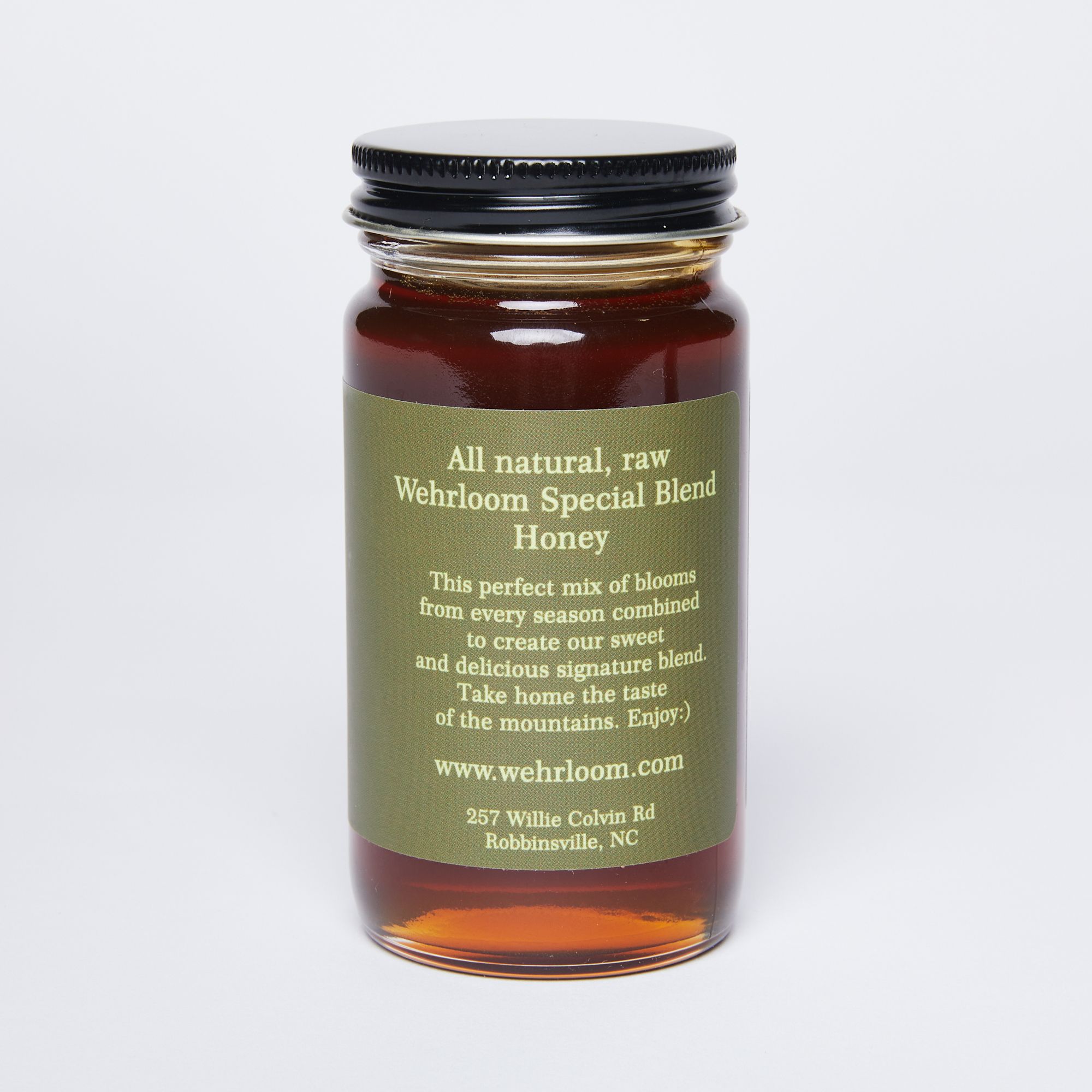 Glass jar filled with golden honey with a black lid and back of green label that reads 'All natural, raw, wherloom special blend honey''.