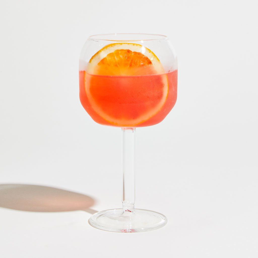 One Velasca calice, a stemmed wine glass with a large, tall bowl, filled 3/4 with a pink liquid with a slice of orange