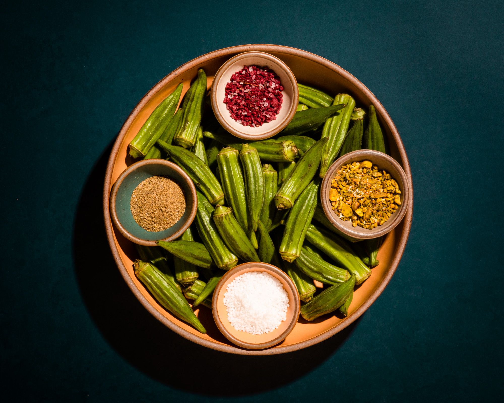 A large shallow serving bowl filled whole okra and 4 small colorful bowls with various colorful seasonings.