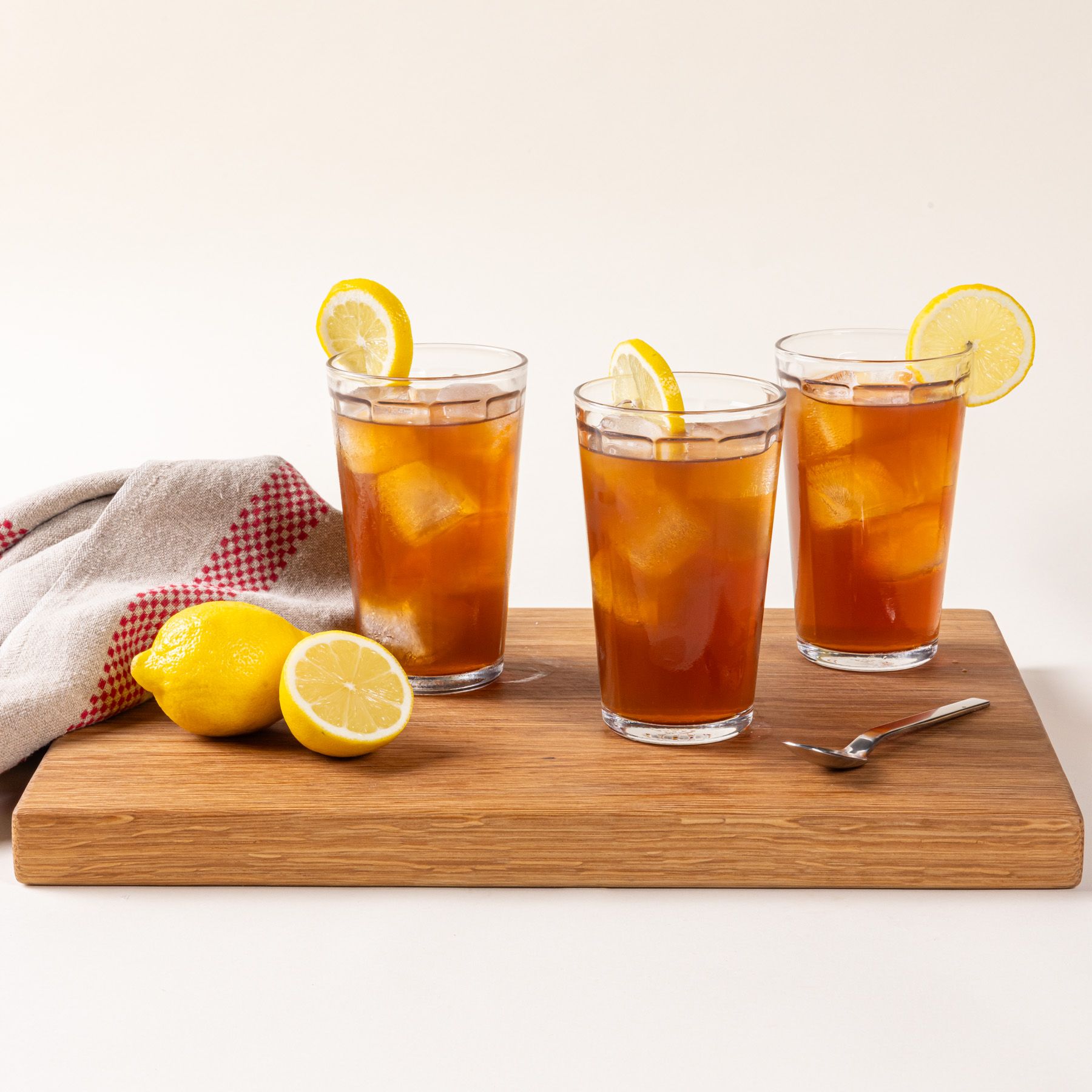 A medium rectangular white oak cutting board with sliced lemons, glasses filled with tea and lemons, a kitchen towel, and a spoon.