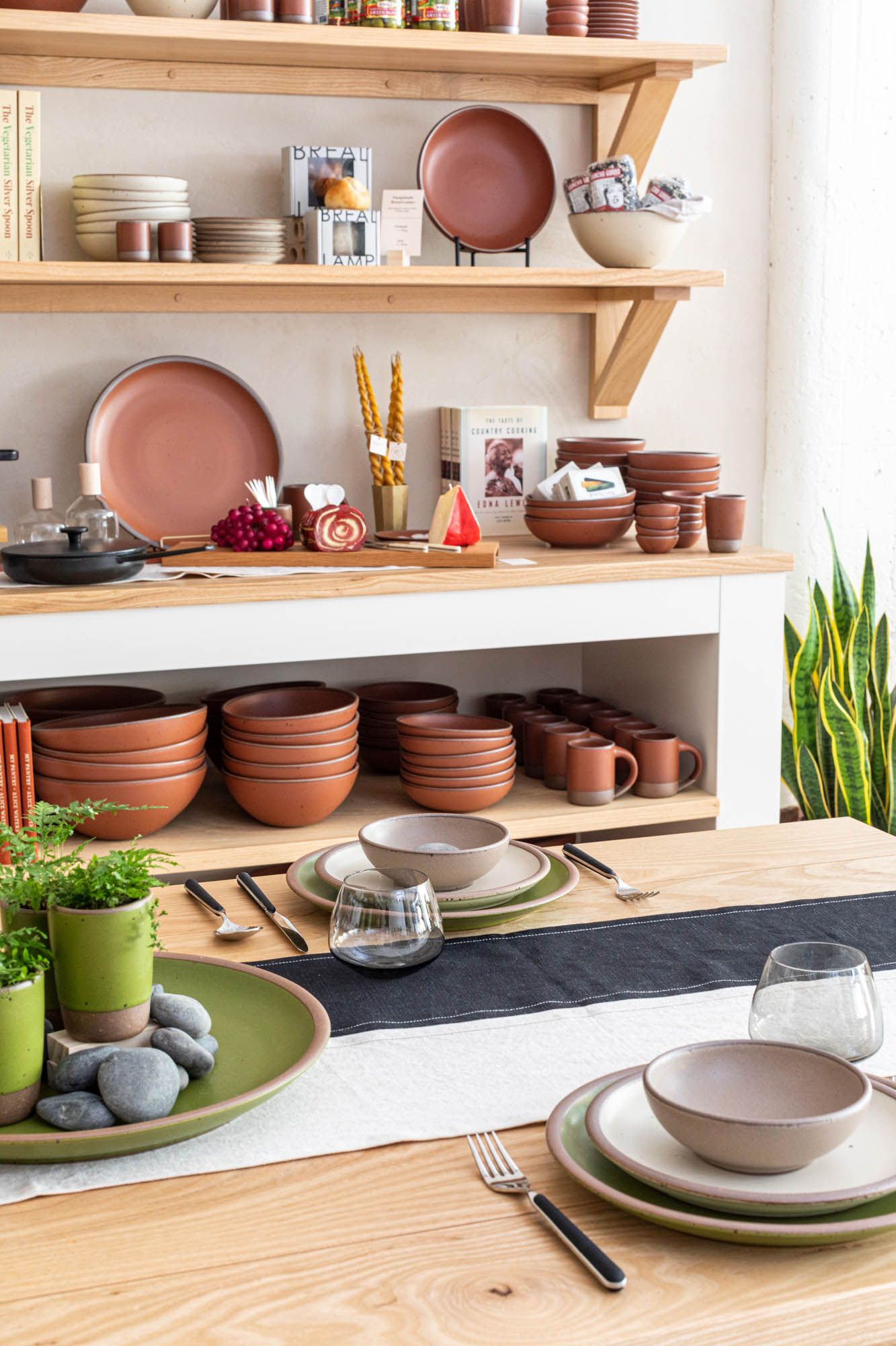 A table featuring ceramic dinnerware place settings with a large display of shelves behind featuring more pottery in a terracotta color as well as other objects for sale.