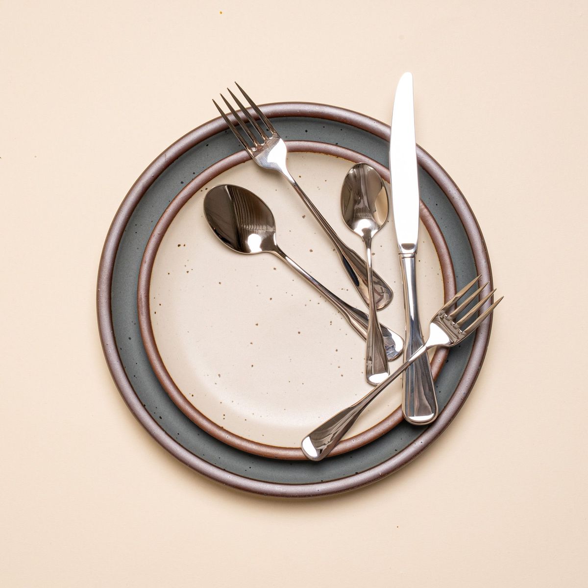 A stainless steel five-piece flatware including 2 forks, 2 spoons, and 1 knife, in a polished finish and classic design. This set is on a place setting with ceramic dinnerware.