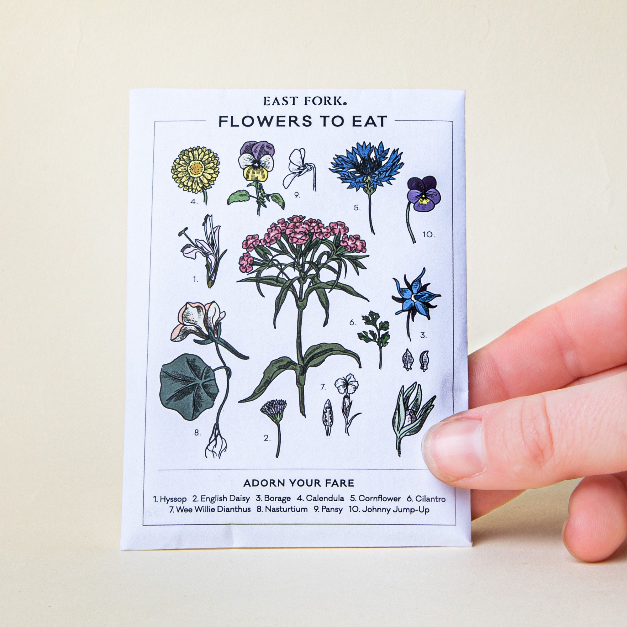 A hand holding a white seed pack that reads "Flowers To Eat" with colorful floral stem illustrations