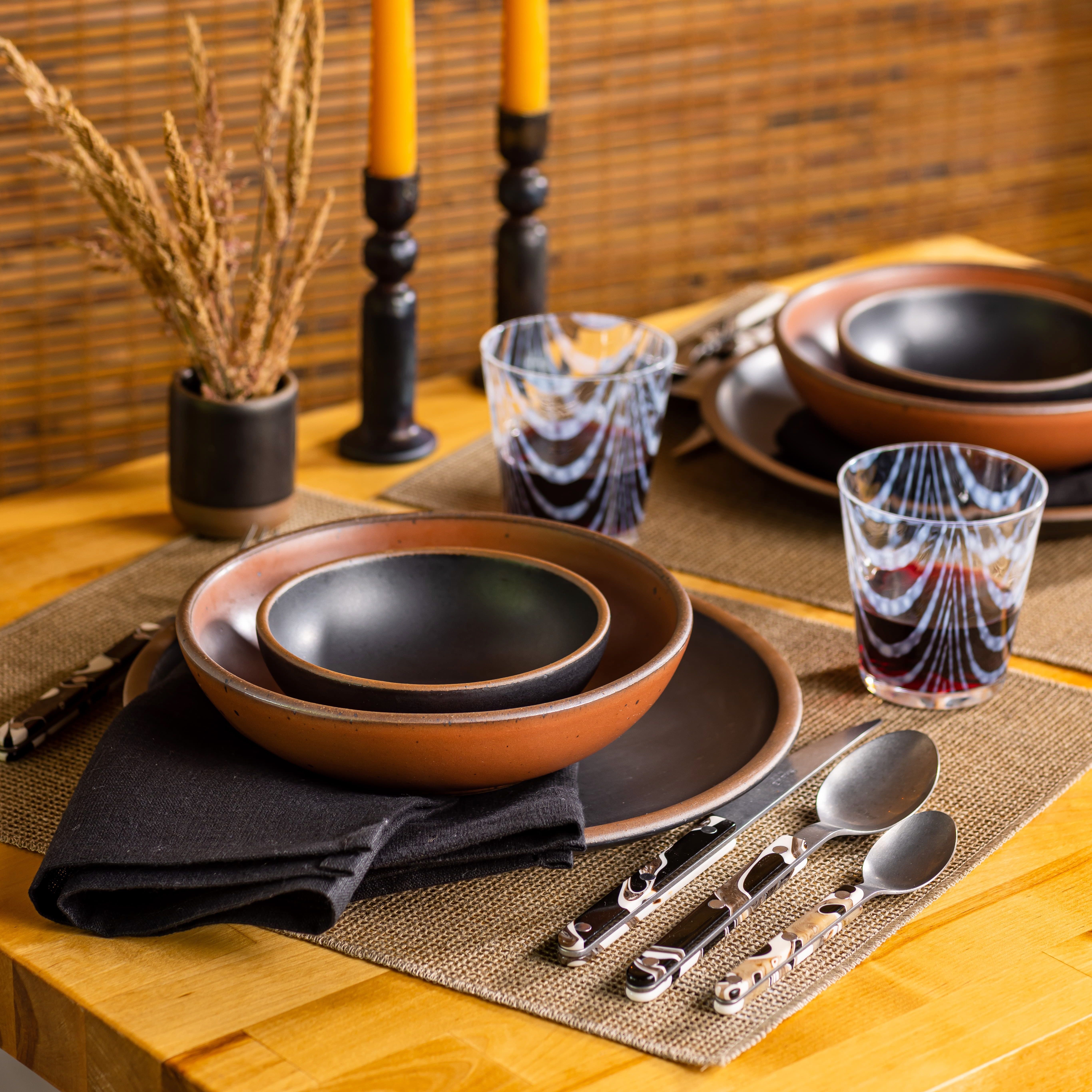 A tablescape with a large terracotta bowl stacked with a smaller black bowl inside. Also on the table are dried plants, flatware, black napkins, natural placemat, and short textured glasses.