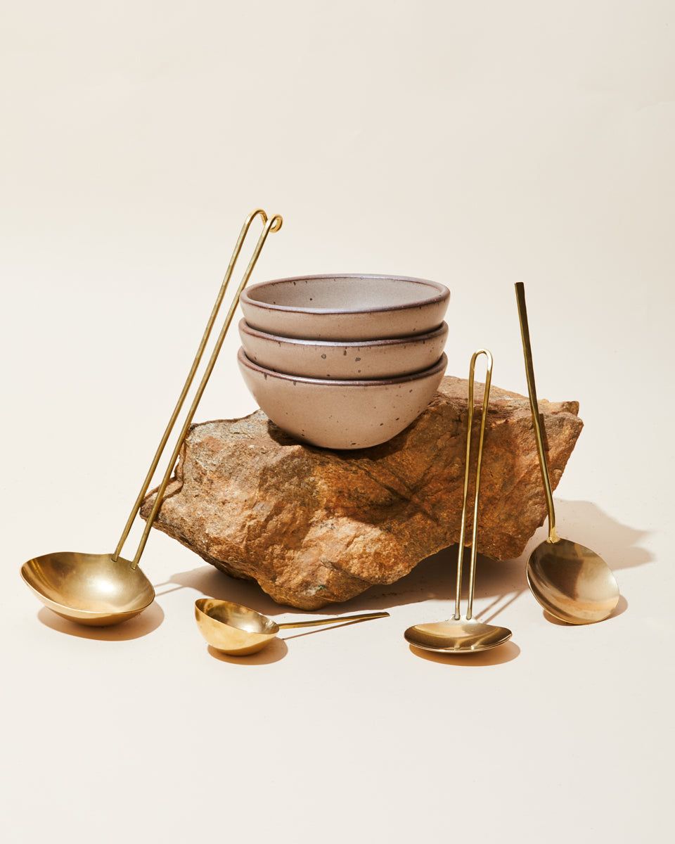 Against a cream background, a stack of ceramic bowls sits on a large rock, surrounded by brass serving utensils of different shapes.