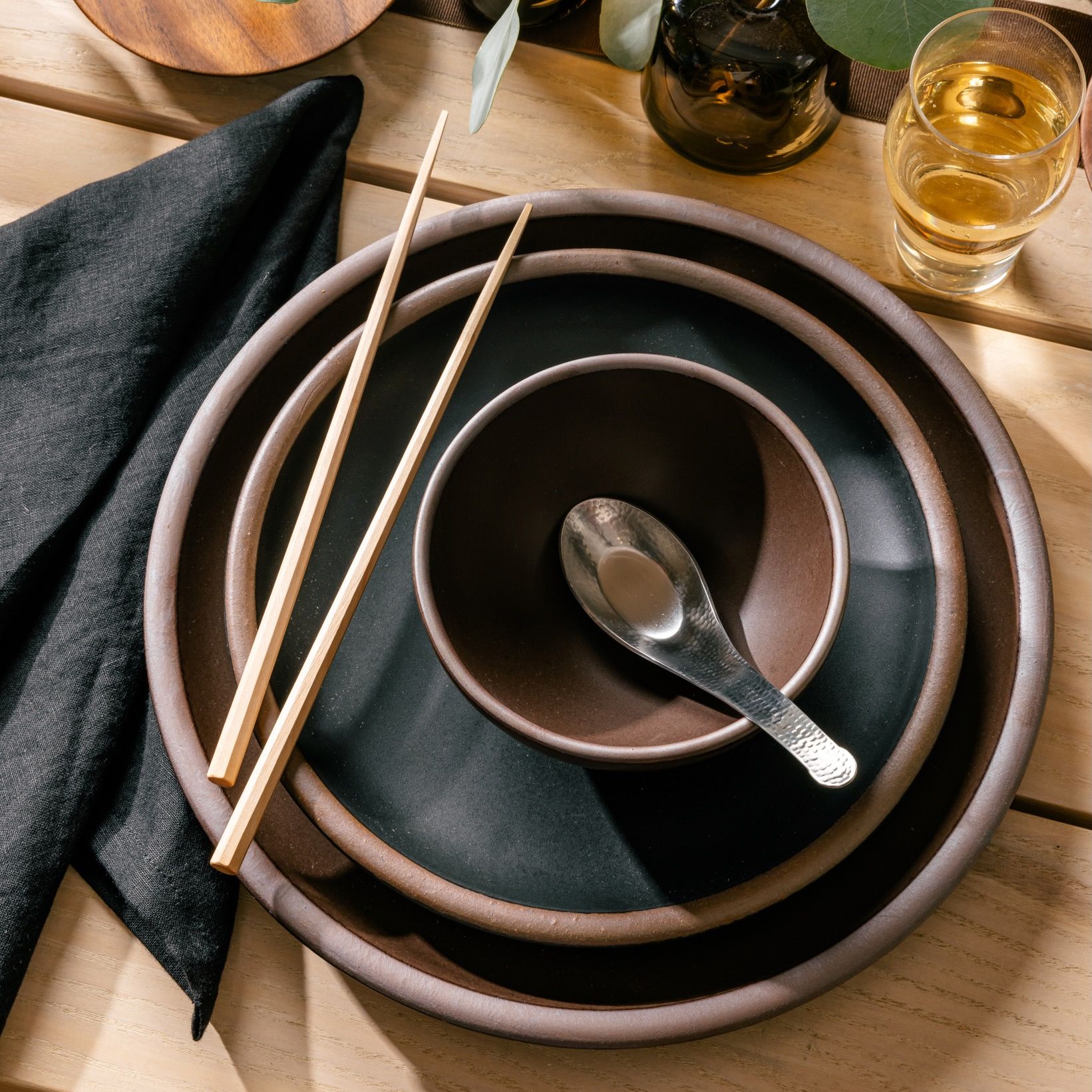 A table setting featuring a dark brown and dark teal large plate, side plate, and small shallow bowl. On top is a hammered steel soup spoon, and wooden chopsticks. Also pictured is a folded black napkin and a clear glass filled with a drink.