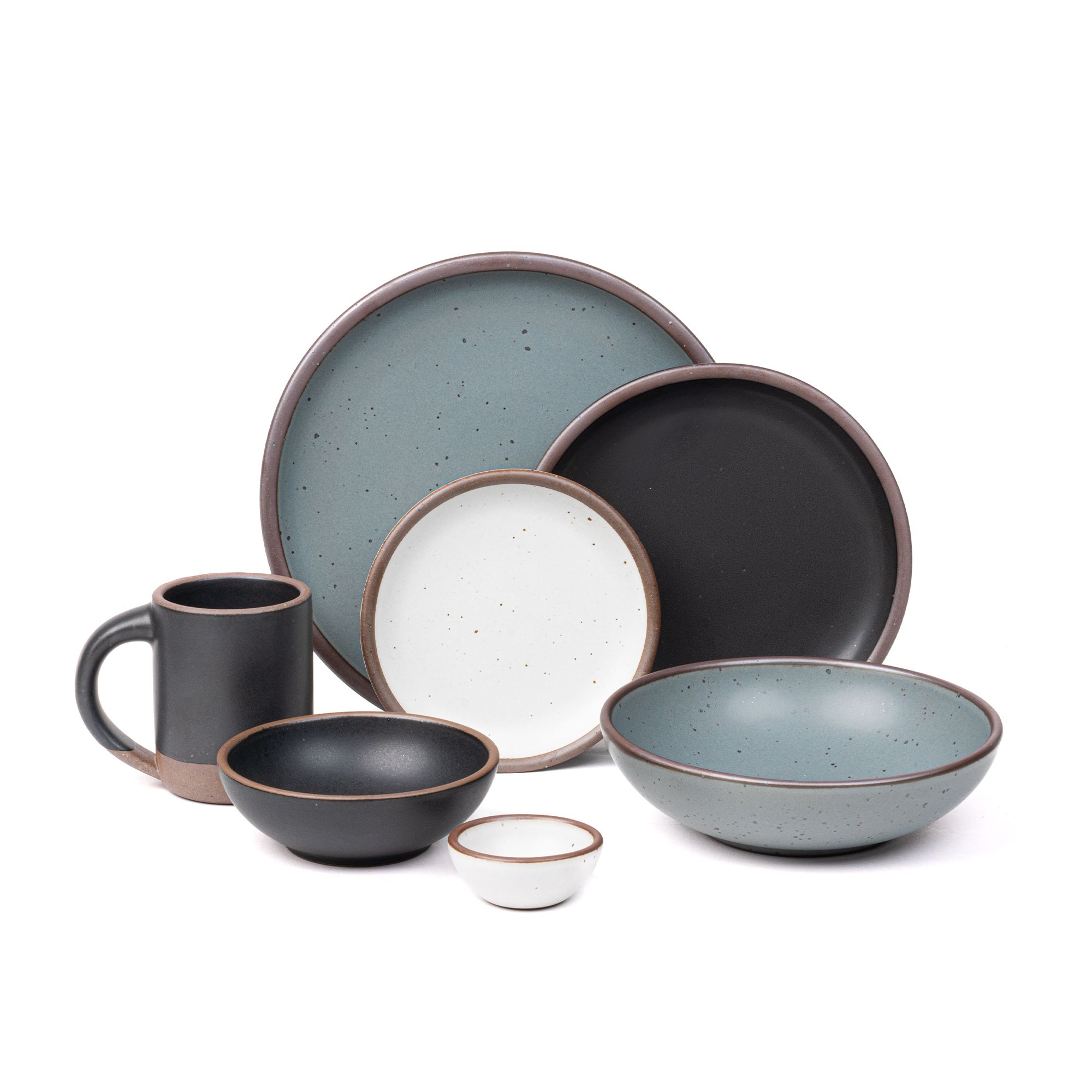 The Mug, side plate, and breakfast bowl in graphite black, the bitty bowl and cake plate in cool white, the everyday bowl and dinner plate in a blue-grey color.