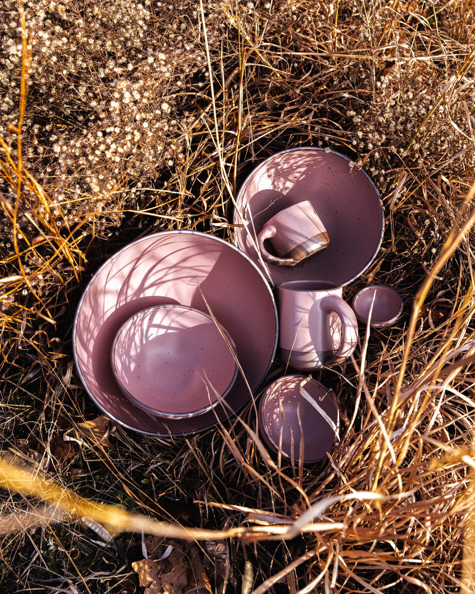 Dusty mauve pink ceramic plates, bowls, and mugs arranged in dry grass and wild plants outdoors.
