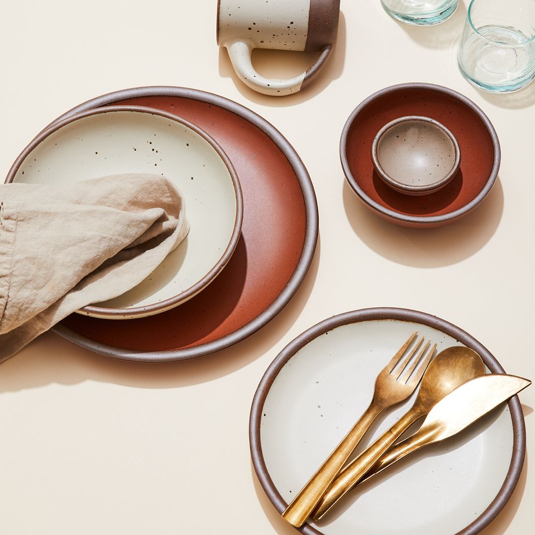 Styled ceramic plates and bowls in cream, white, and dark terracotta colors all close together. A bowl has a folded beige napkin and a plate has a brass fork, spoon, and knife placed on it.