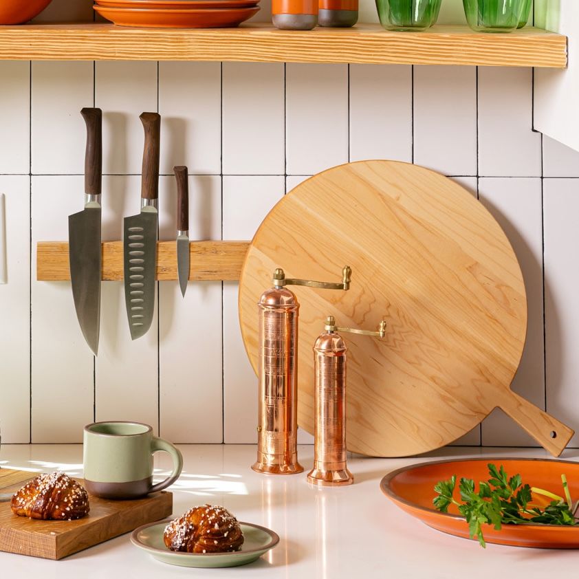 On a kitchen counter, is a stylish prop set up with a knife block on the wall with 3 kitchen knives, a copper pepper and salt mill, a round wooden serving board. In front of all of these items are ceramic plates filled with ingredients and morning buns, and a short ceramic mug in sage green.